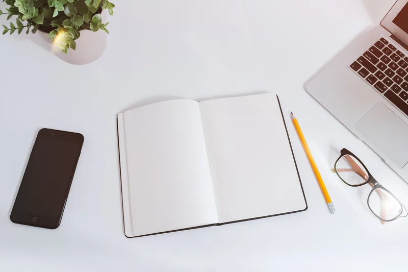 A blank notebook, smartphone, pencil, glasses, and laptop arranged neatly on a white office desk.