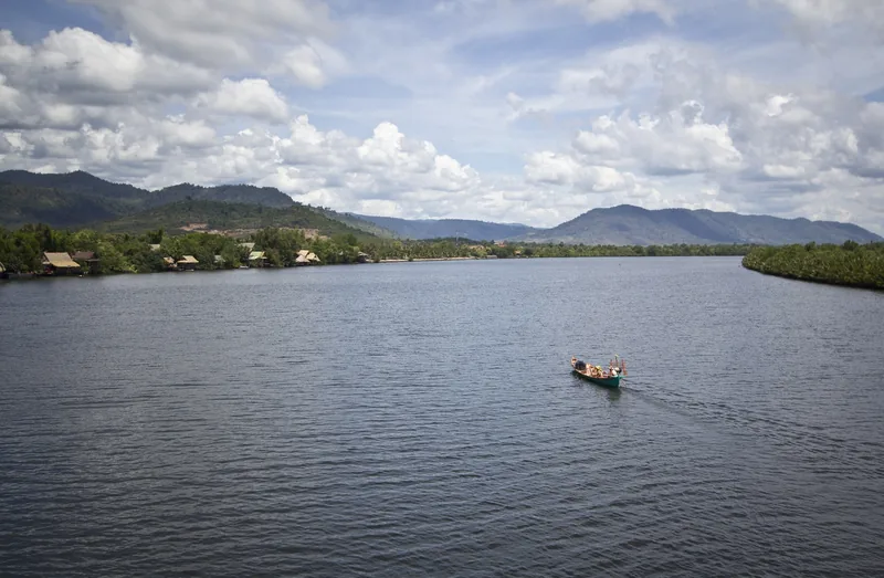 A small boat travels upstream on the Kampot River, with mountains and stilt houses in the distance.