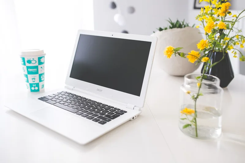 A white laptop sits on a desk beside a coffee cup and yellow flowers in a bright home office.