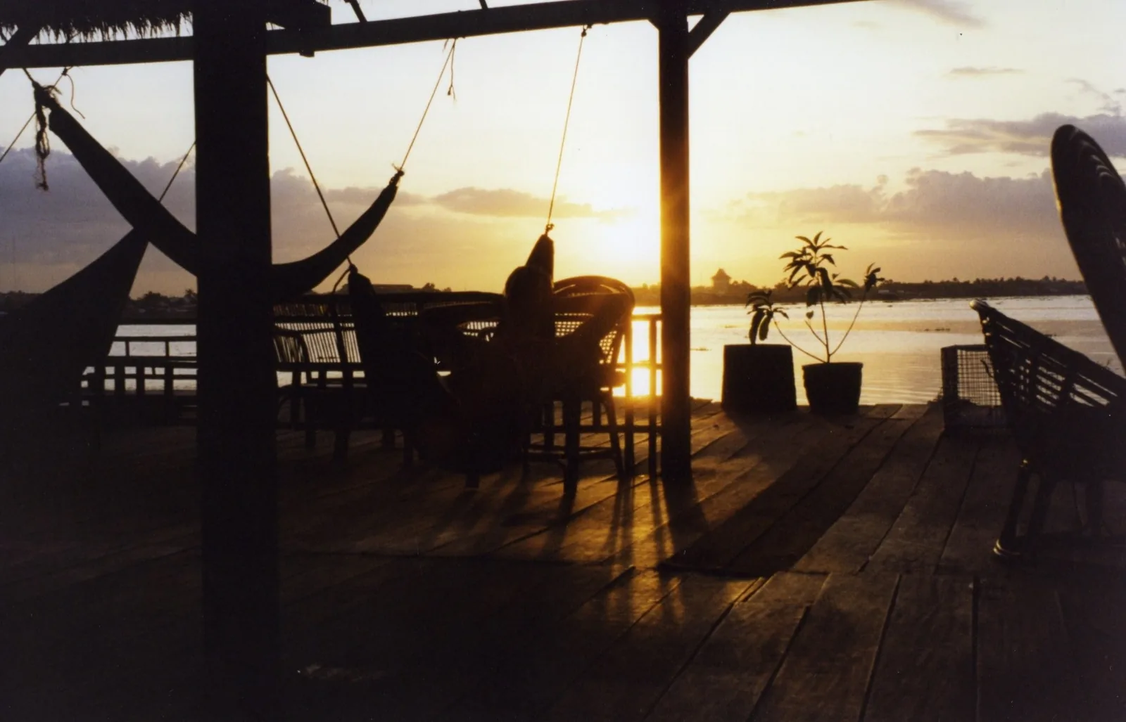 Sunset silhouettes a wooden riverside deck with hammocks and chairs at a guesthouse in Phnom Penh.