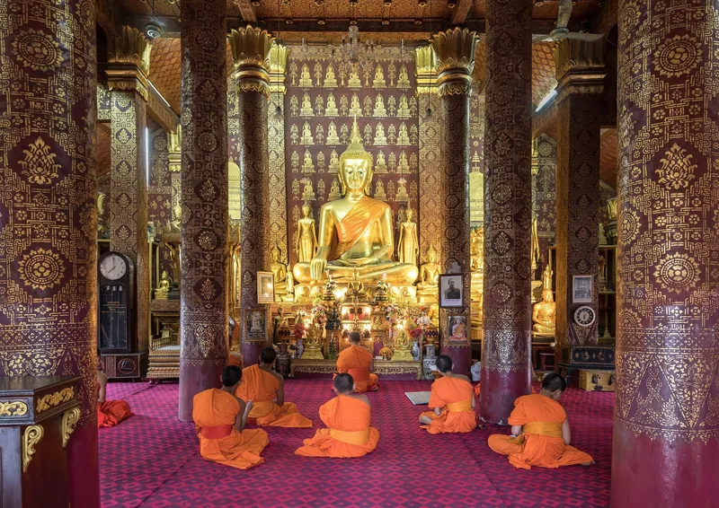 Buddhist monks in orange robes pray before a golden Buddha statue inside a temple in Luang Prabang.