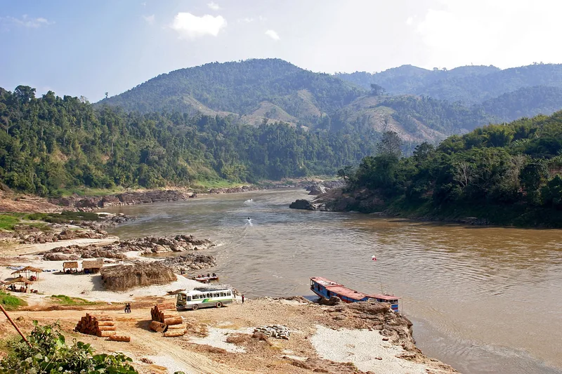 A river confluence near Xieng Kok in Luang Namtha, with a bus and boats waiting on the sandy shore beneath forested hills.