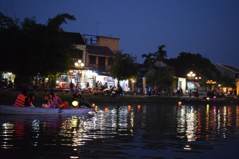 A wooden boat with passengers in life jackets floats on the river past floating candles and riverside cafes in Hoi An at.