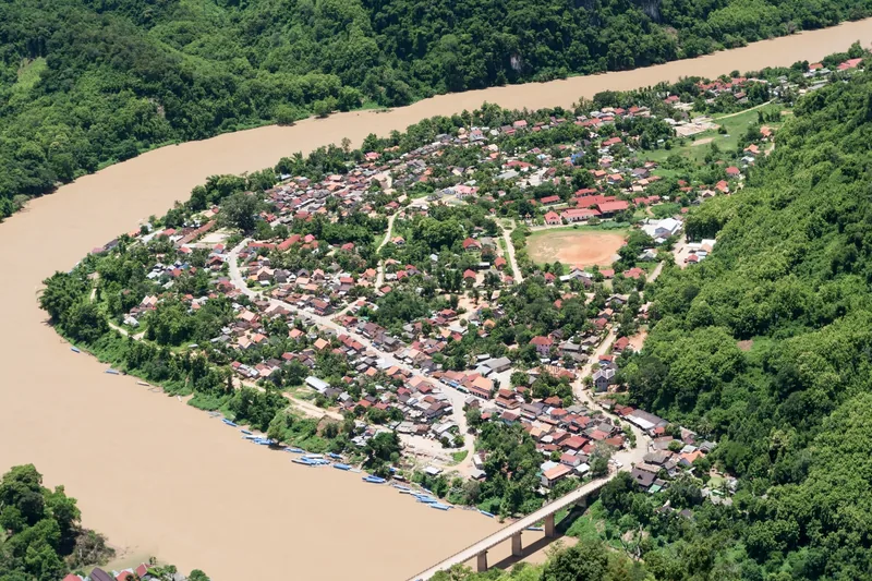 Aerial view of Nong Khiaw village on a bend of the muddy Nam Ou River, surrounded by forested limestone hills.